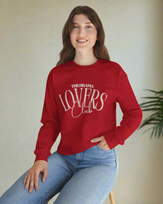 Person wearing a red sweatshirt with 'The Drama Lovers Club' text, sitting on a white surface with a plant in the background.