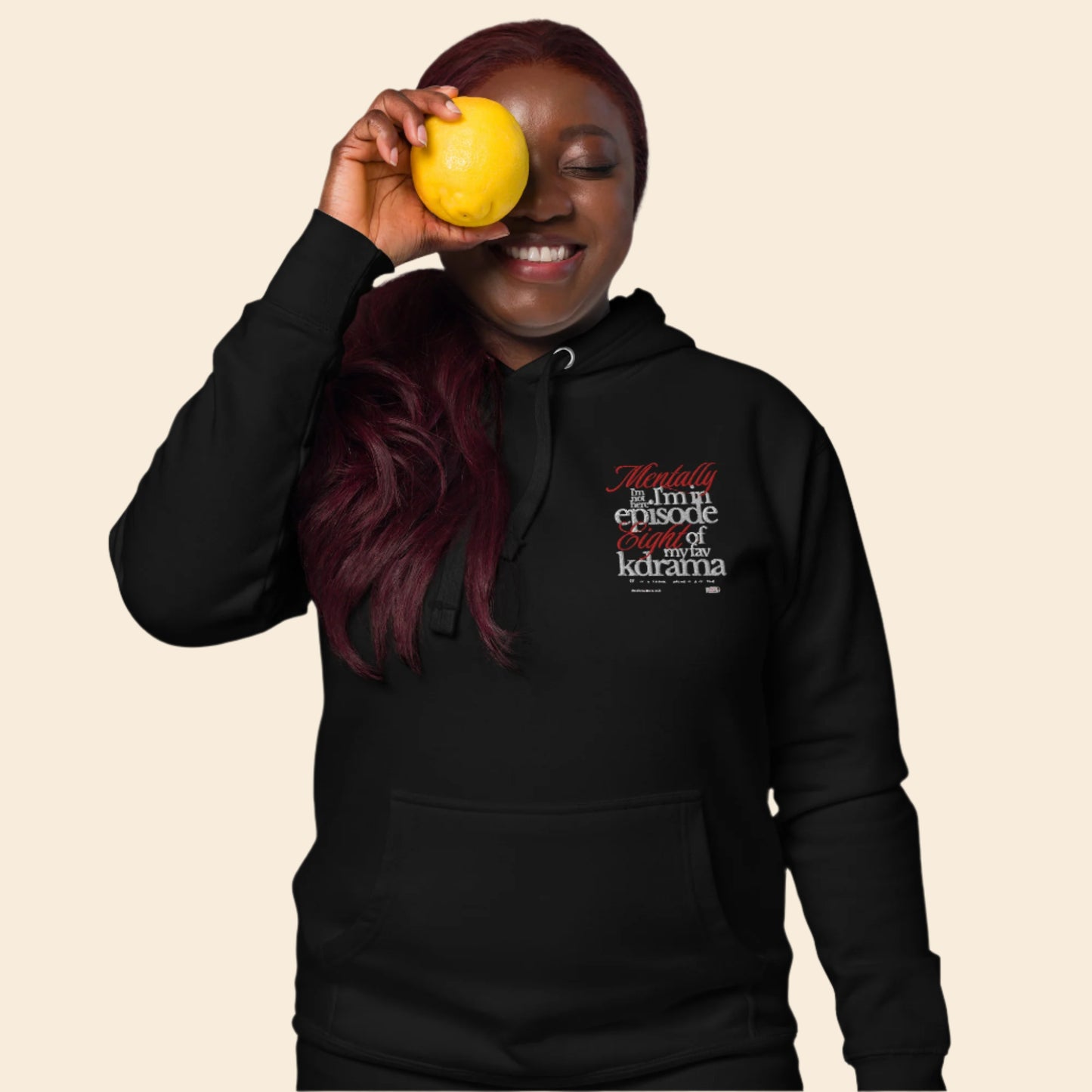 Person wearing a black hoodie with text, mentally I am on episode 8 hoodie holding a yellow fruit against their eye.