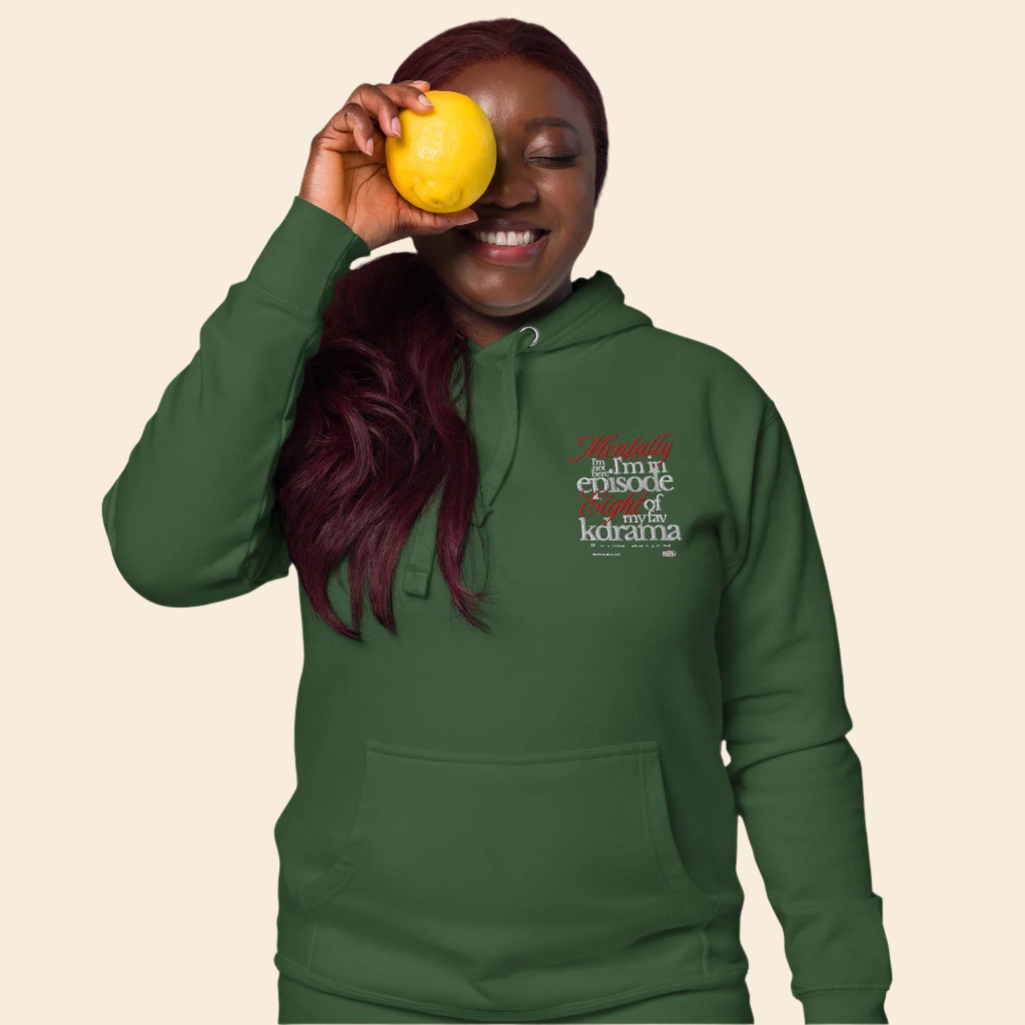 Person wearing a green hoodie with text mentally I am on episode 8 hoodie, holding a yellow fruit against their eye.