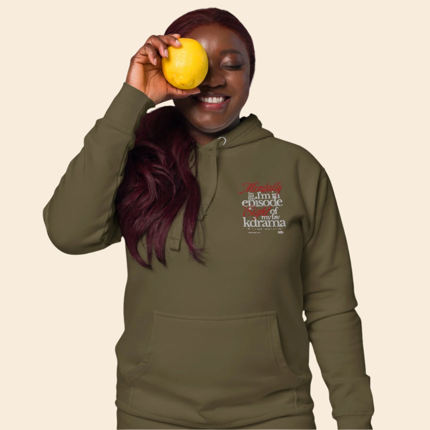 Person wearing a green hoodie with text mentally I am on episode 8 hoodie, holding a yellow fruit against a white background