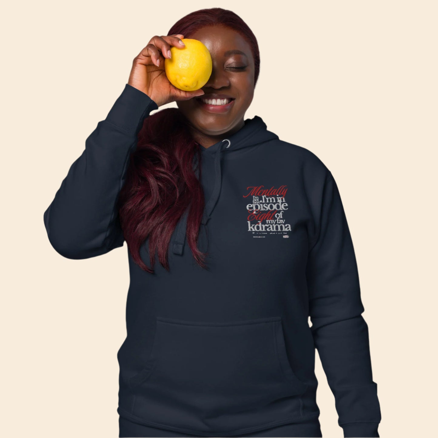Person wearing a navy hoodie with text mentally I am on episode 8 hoodie, holding a yellow fruit against their eye.