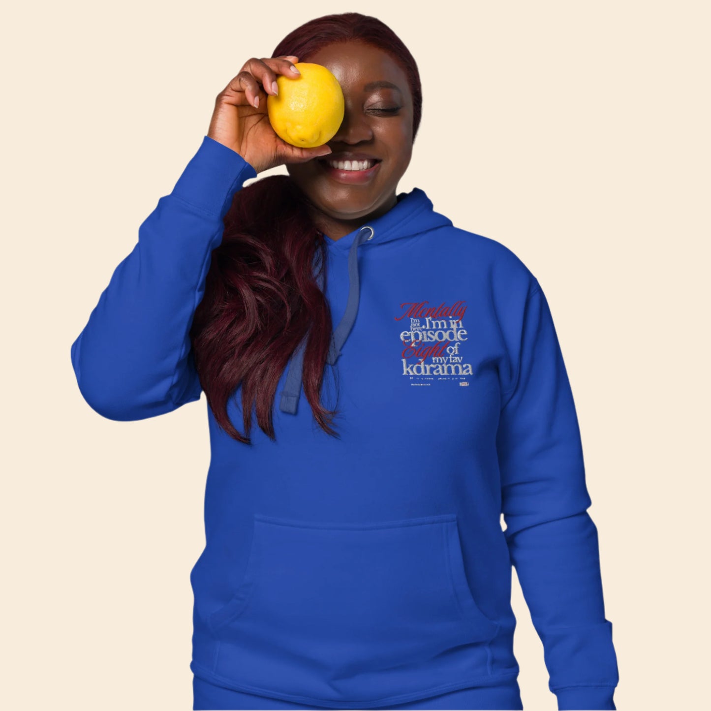 Person wearing a blue hoodie with text mentally I am on episode 8 hoodie, holding a yellow fruit against their eye.