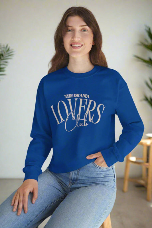 Person wearing a blue sweatshirt with 'The Drama Lovers Club' text, sitting on a white surface with a plant in the background.